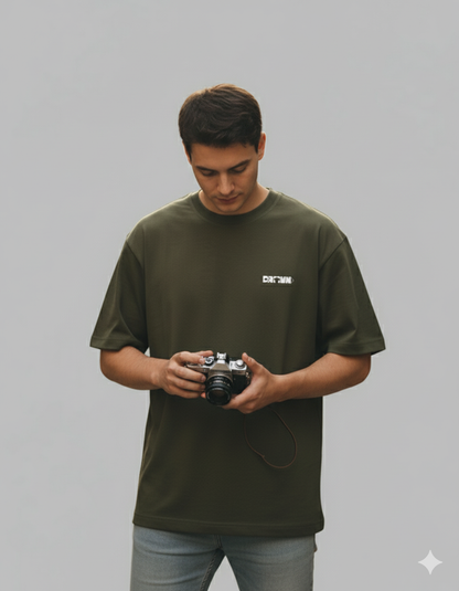 Man holding a camera wearing an olive green t-shirt with a logo on a gray background