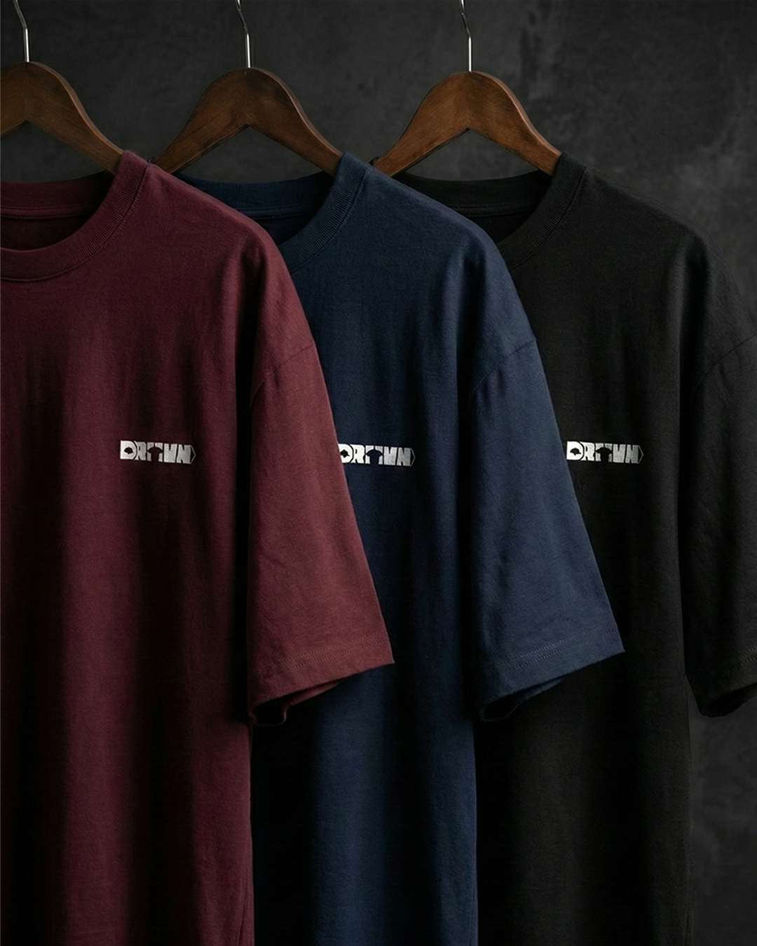 Three t-shirts in maroon, navy, and black hanging on wooden hangers against a dark background.